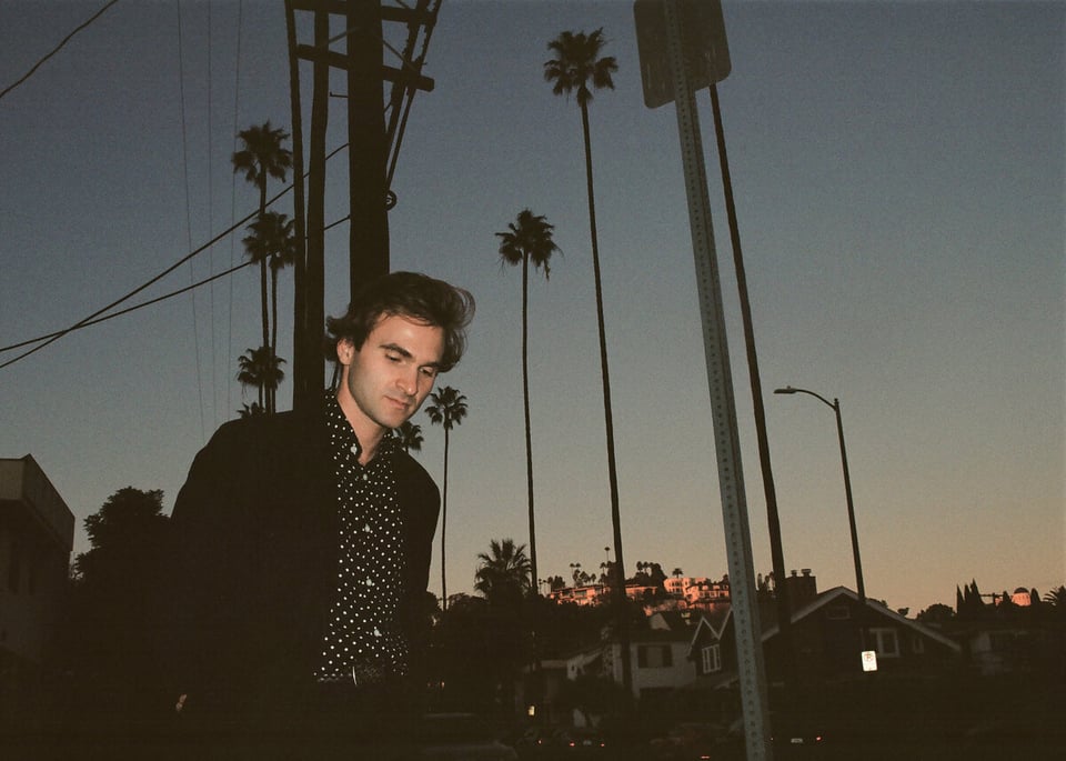 A man in a dark sport coat and black button-down shirt with white dots walks down a street at dusk, with towering palm trees in the background.