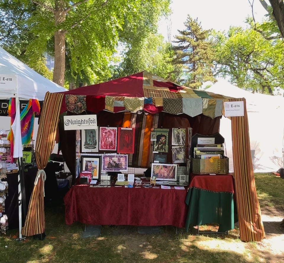 Photograph of an art booth set up. There is a tent with a renaissance faire style tent, and prints set up on display walls. There is a treasure chest full of prints. The overall effect is very fantasy medieval.