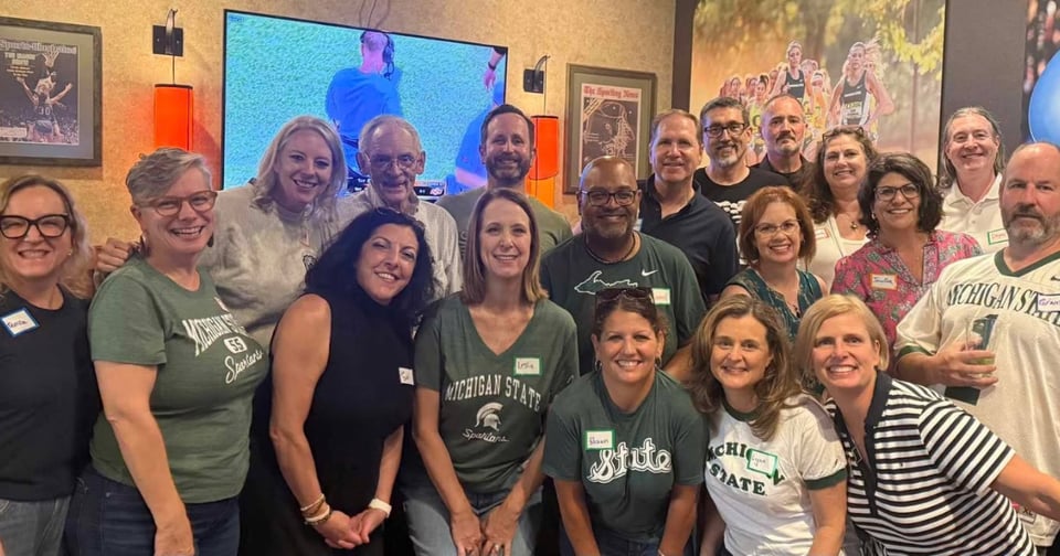 A group of people, many wearing Michigan State Spartans apparel, are posing together indoors. They appear to be at a social event or reunion, with sports memorabilia and framed pictures visible in the background.
