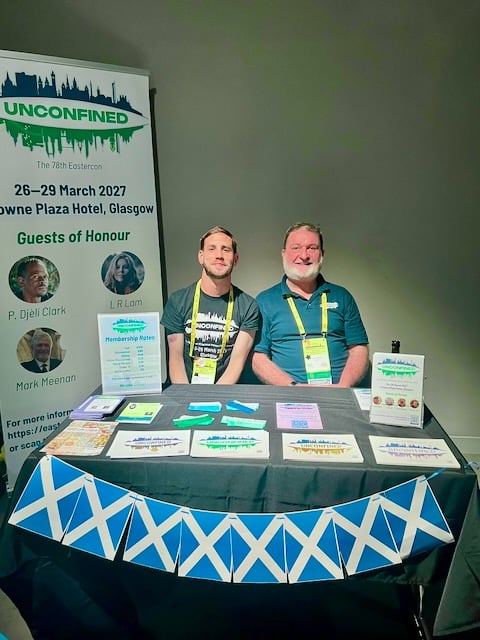 Two people sitting at a table covered in promotional material for the convention.