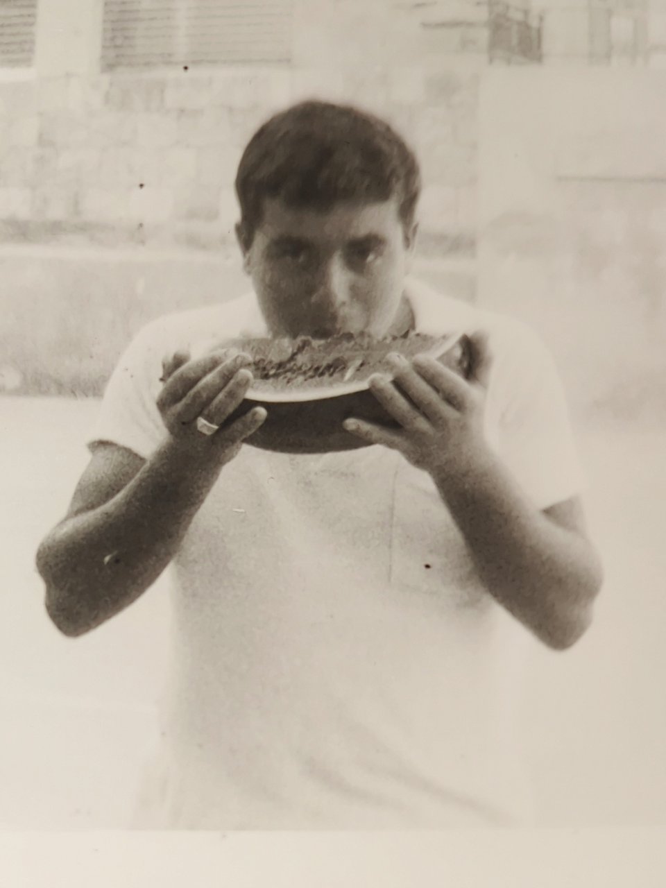 A black and white photo of a young man with black hair wearing a white t-shirt and holding up a slide of watermelon to his mouth with both hands.
