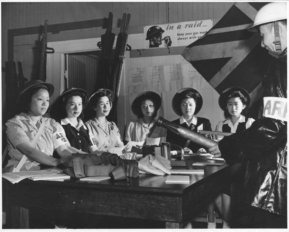 Black and white photo. A group of women wearing helmets and ARP arm bands listen to a man demonstrating something with a model of a small bomb.