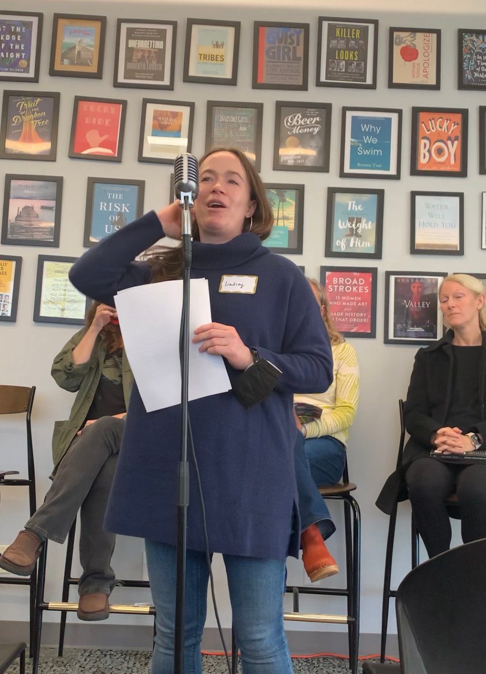 The image shows the author standing at a microphone, smiling and holding sheets of paper, in a long blue sweater and blue jeans. Three people are seated behind her, and on the wall behind them are covers of books.