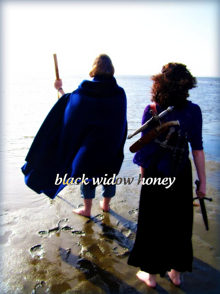 two figures viewed from behind as they face out toward the sea; the front figure is taller and appears older, wearing a long cloak and holding a staff. the second figure is smaller and female-presenting, and carries six daggers of various size and shape.