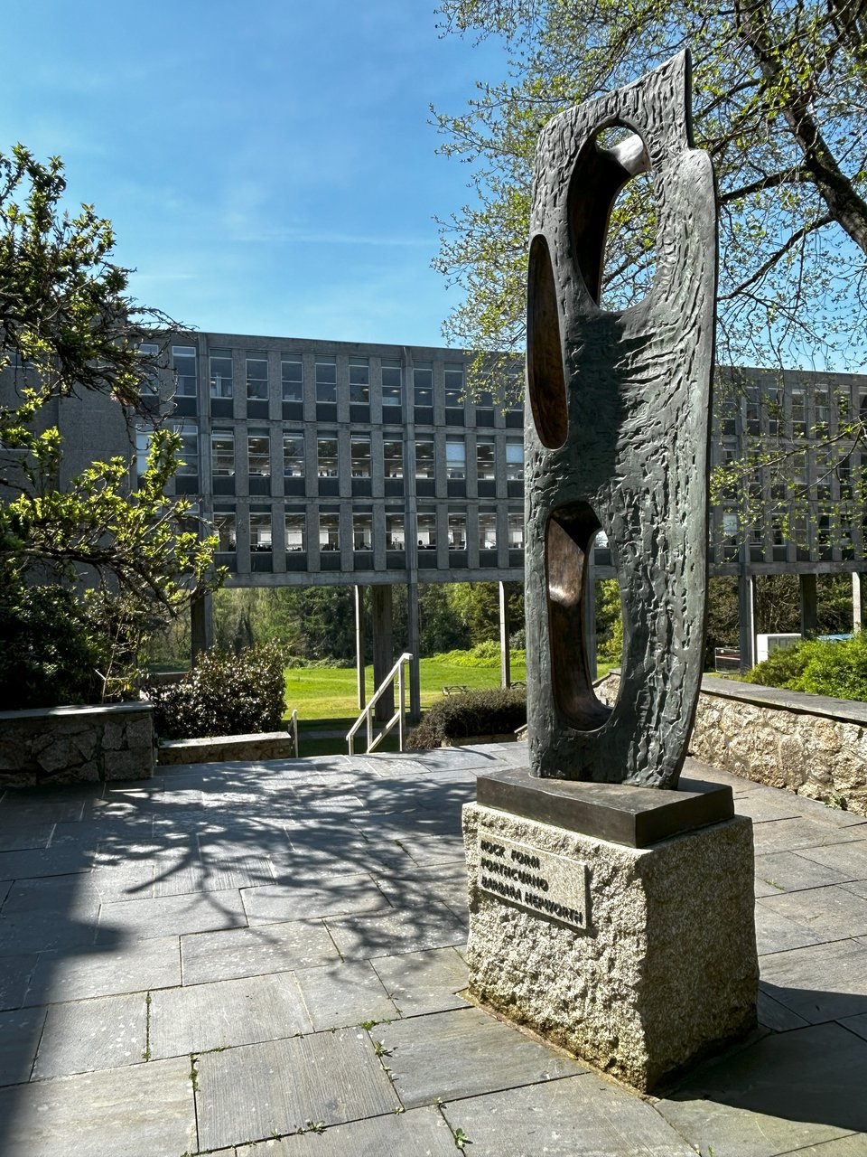 the sculpture is a bronze monolith with curved edges. It's been pierced in several places, revealing smoother interior surfaces. It's bigger than a person and sits on a plinth in a courtyard. Beyond it, steps drop down to the raised section of the building, and the green lawns beyond are visible. Within the courtyard, mature trees obscure some of the building.