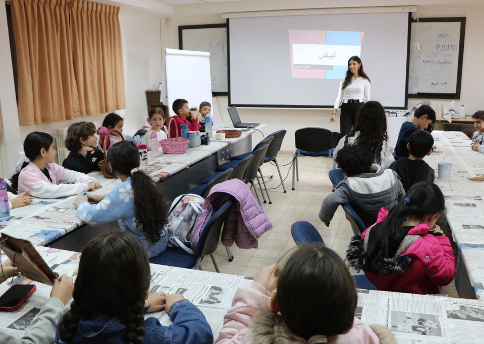 Chidlren sitting in a U-shape at tables, with a teacher at the front of the room in front of a slide with one word written in Arabic