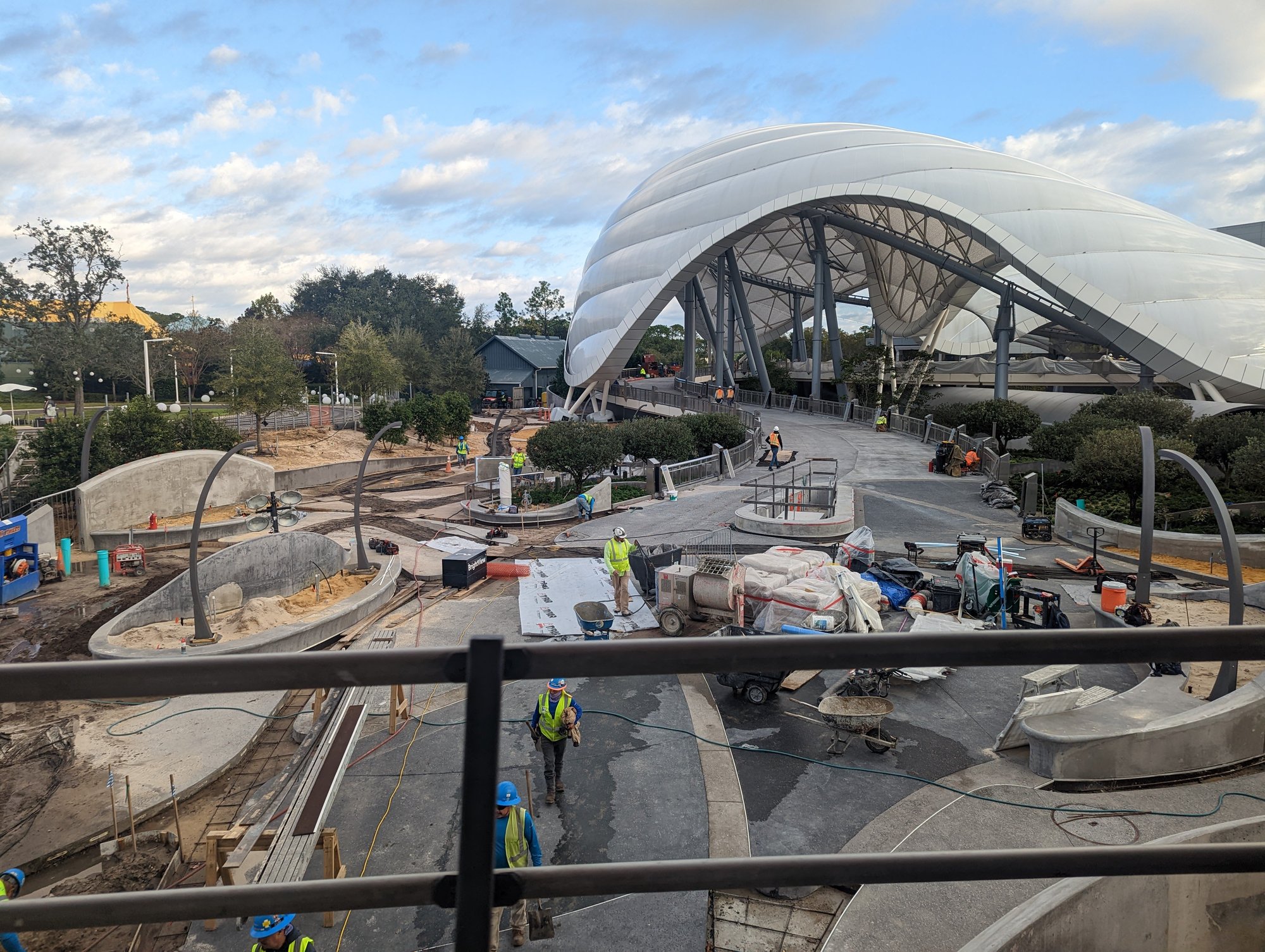 The view from the PeopleMover of the Tron roller coaster under construction. The wavy white canopy dominates the scene, and construction workers work on newly-paved concrete paths below