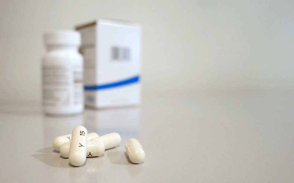 Close-up image of several white pills on a smooth, reflective surface. Some of the capsules are marked with black text ("Y 15"). In the blurred background, there is a white prescription pill bottle and a pharmaceutical box with a blue stripe, suggesting a clinical or medical context. The image evokes themes of healthcare, medication, or pharmaceutical treatment.
