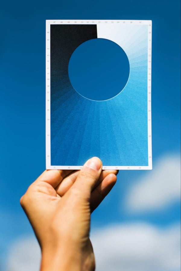 A white hand holds up a postcard with a round cut out in the center and various shades of blue surrounding it. the blue sky shines through the middle.