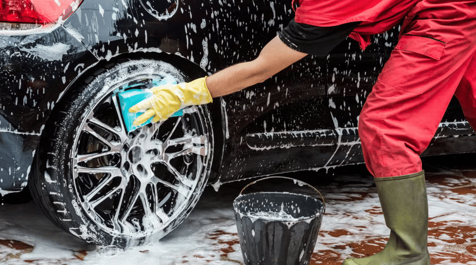 A person wearing a red jumpsuit and green rubber boots cleaning a car with a big sponge.