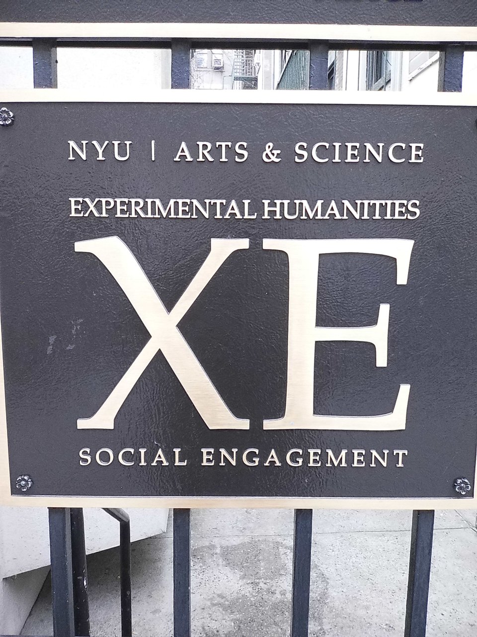 Digital Instax photo of a sign on a fence at NYU for Experimental Humanities Social Engagement, with a large XE at the center