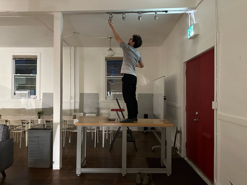 An absurdly handsome man changes a lightbulb while standing on a table.