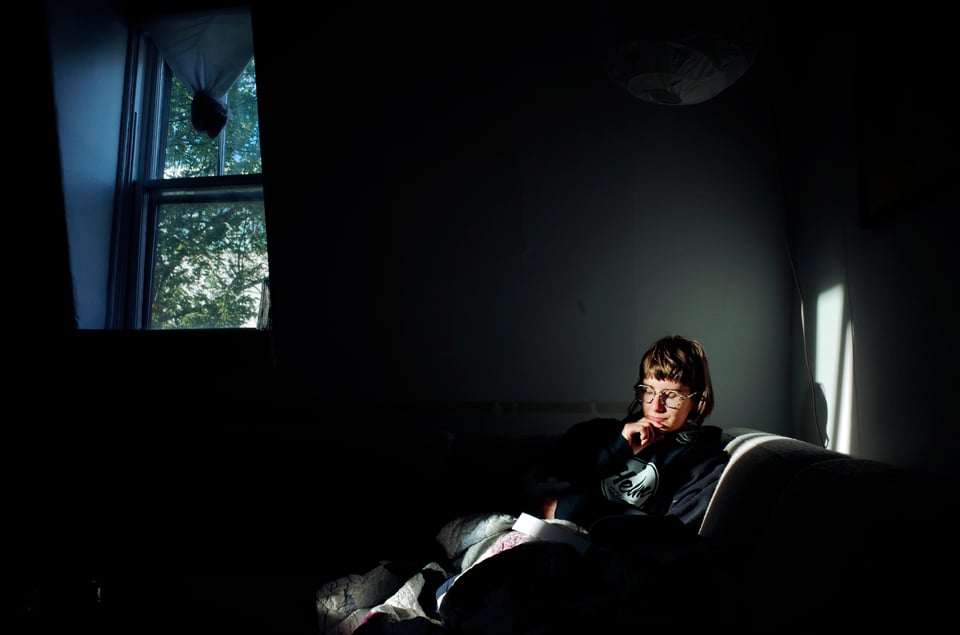 A picture of Sophie reading on their couch in a beam of morning sunlight. Leafy trees can be seen outside the window. The picture of otherwise dark.