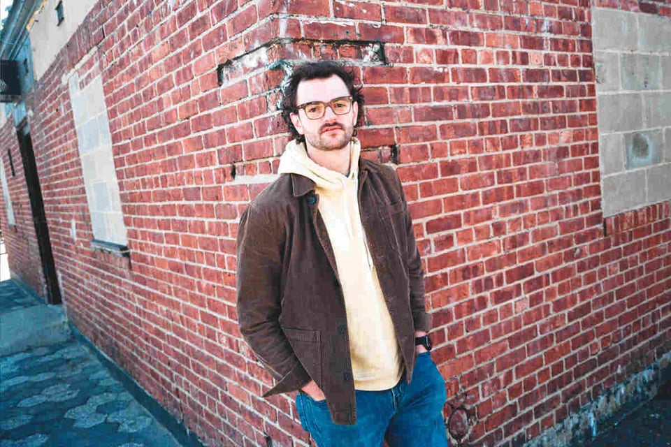 A man with glasses and a faint beard stands at the corner of a brick wall, wearing an off-white hoodie, a brown jacket and blue jeans.