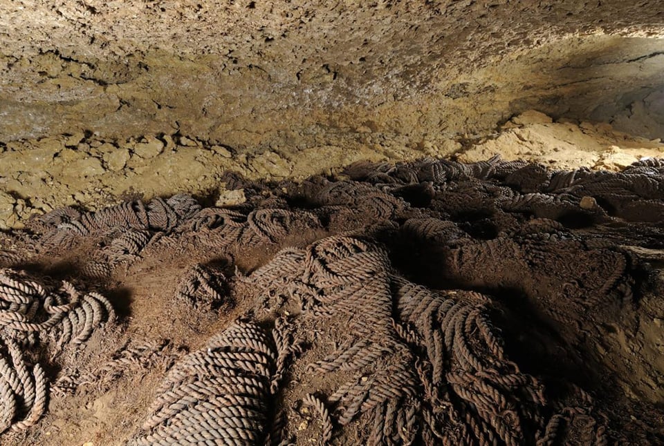 A cave floor is covered in thick, twisted lava formations that resemble coiled ropes, their surfaces dark and textured. The ceiling above is rough and pale, lit to reveal layers of rock and mineral deposits. The scene captures the striking natural patterns of hardened lava, frozen mid-flow in intricate, looping shapes.