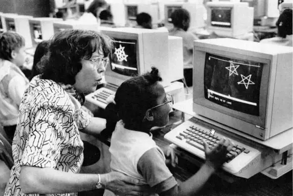 black and white photo of a young Black girl learning Logo on a Apple computer. a white teacher looks over her shoulder