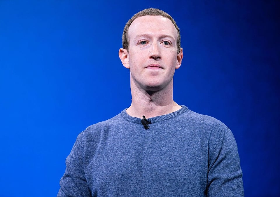 A white man stands against a gradient blue background wearing a blueish grey pullover, with a small mic at his neck. His lips are pursed and he is staring intensely.