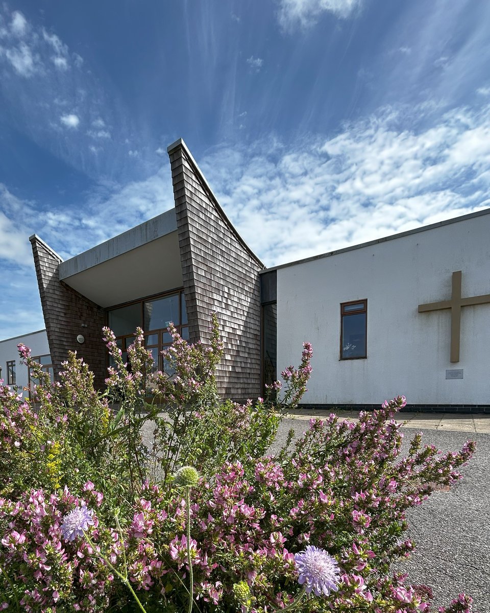 A modern church. It’s a single storey with white render walls apart from in the middle where two cedar shingle clad walls rise up like wings. There’s wildflowers in the foreground.
