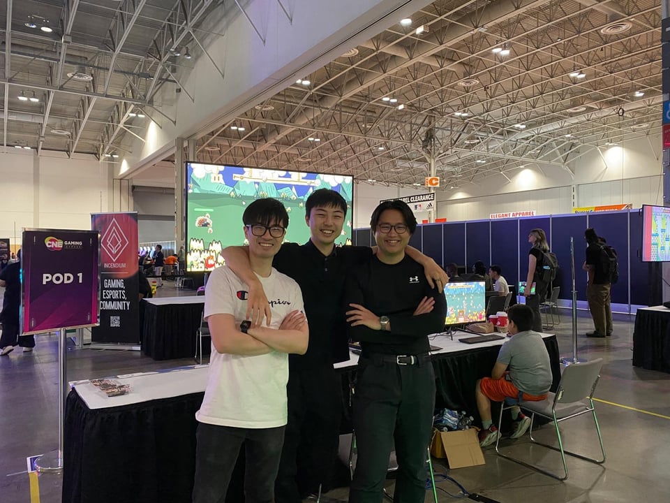Three young east-asian men stand in front of their booth at a gaming conference. The one in the middle has his arms around the others. They all have short black hair and the two on the outside are wearing glasses.