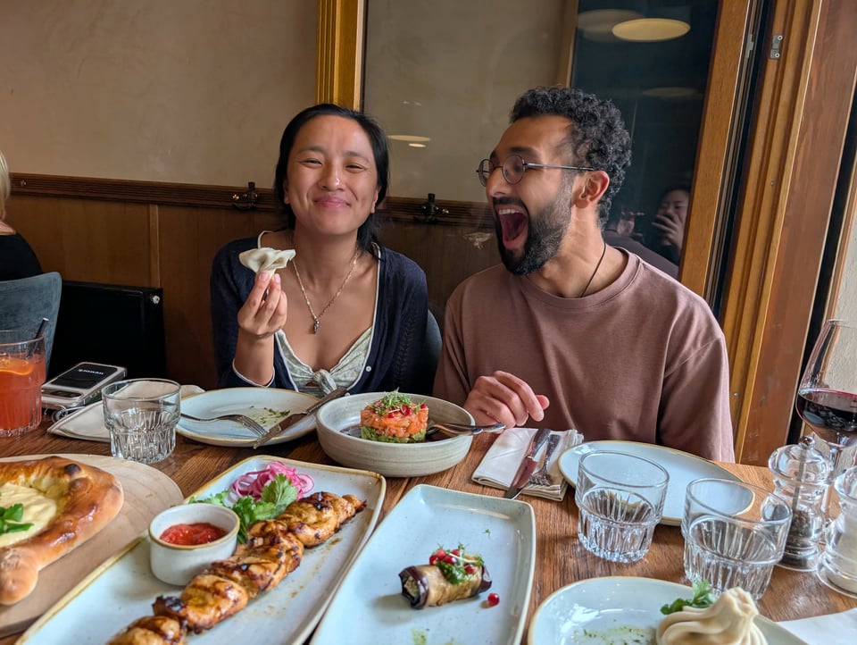 Dillan and Donna smiling in front of a table of food, Donna holding a large soup dumpling and DIllan pretending to eat it.