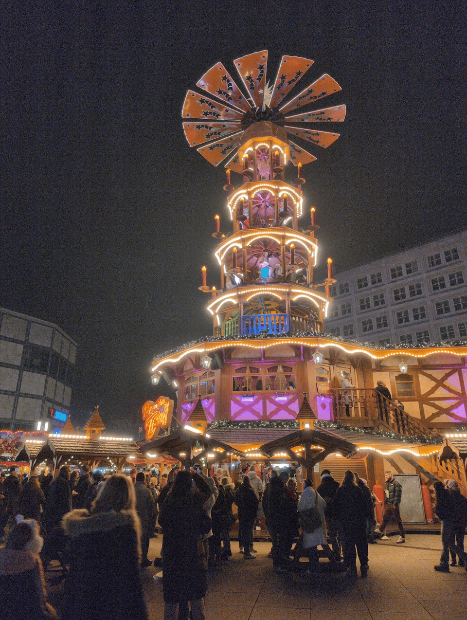 A life-sized Christmas pyramid stands on a market square. There are standing tables with people at them. There are also quaint huts that sell food and items. A Christmas pyramid is a stack of round or octagonal layers that each have Christmas decor. The uppermost levels have candles whose heat makes the fan on top spin. (This one is electronic, though.)