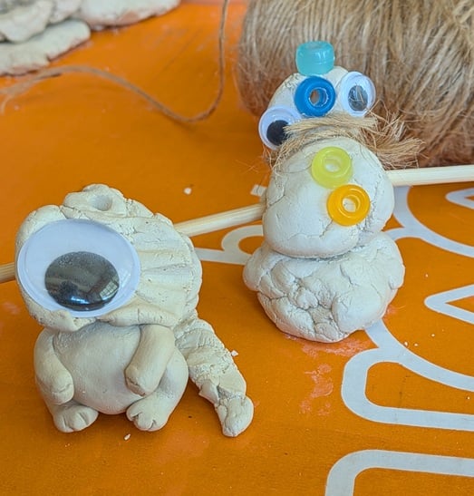 two little creatures made out of white clay; one with four paws sits on its back legs, and has a long lumpy tail and a flat lumpy head with one gigantic googley eye stuck to it. The other looks like a 3 tiered snowman with askew googley eyes, four pony beads (one like a hat, one like a nose, and two as buttons), and a mouth or perhaps scarf made out of twine. They both look simultaneously forlorn and cursed.