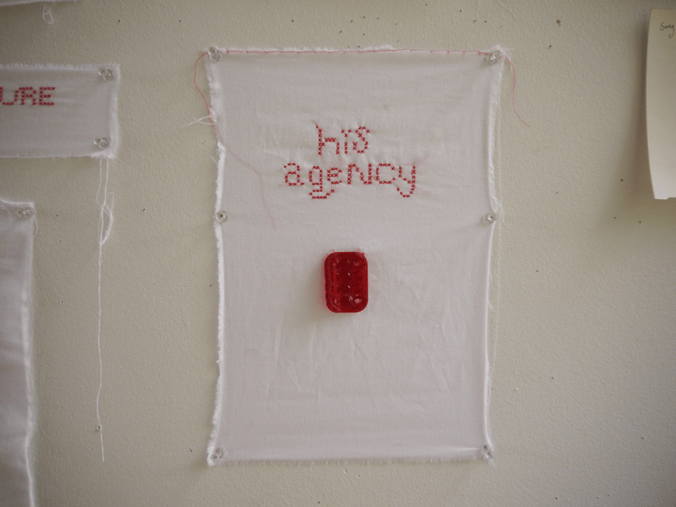 a page of light white cloth hung directly on a wall. in red thread, the words "his agency" are embroidered with small cross stitches. below the words, a plastic reflector is sewn to the cloth.