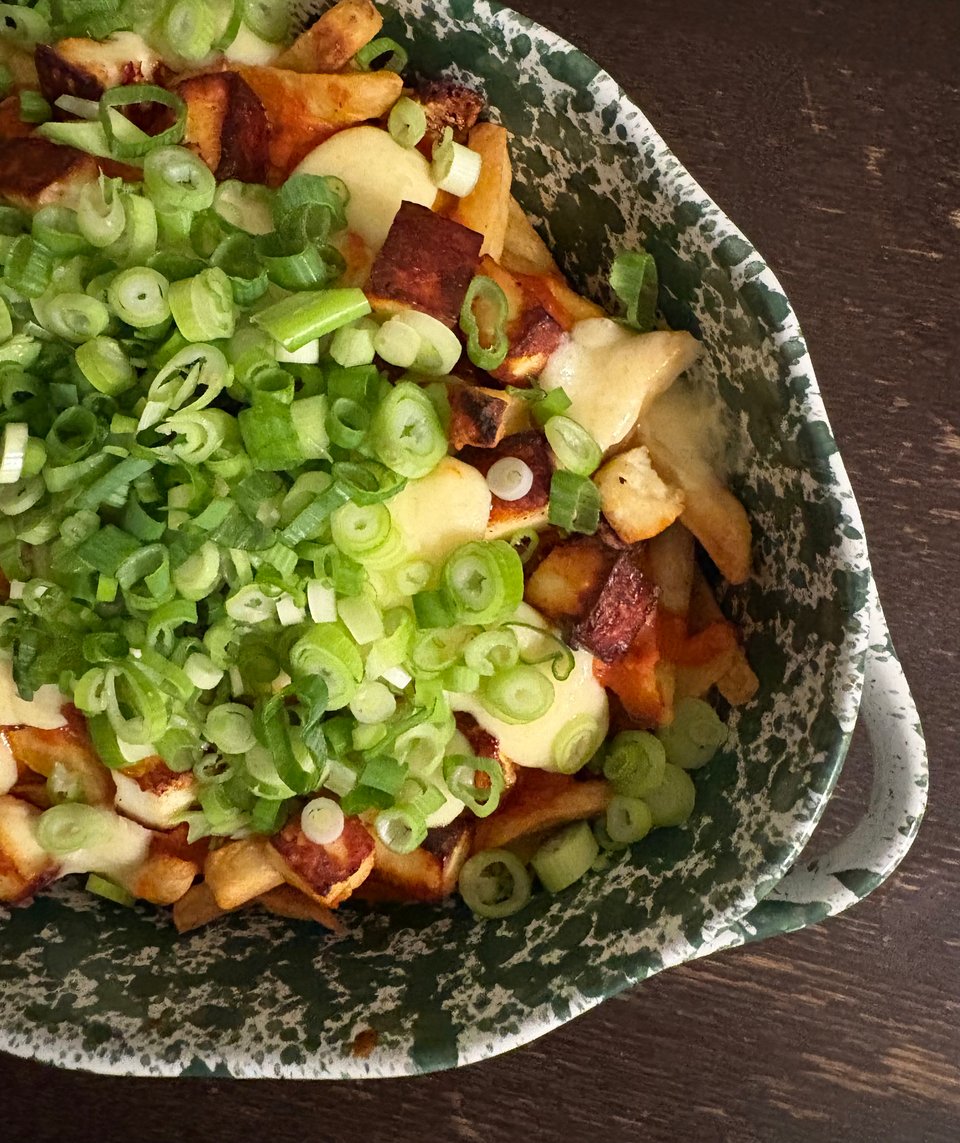 A green and white tray filled with layers of French fries, paneer, cheese curds, masala gravy, and sliced green onions.