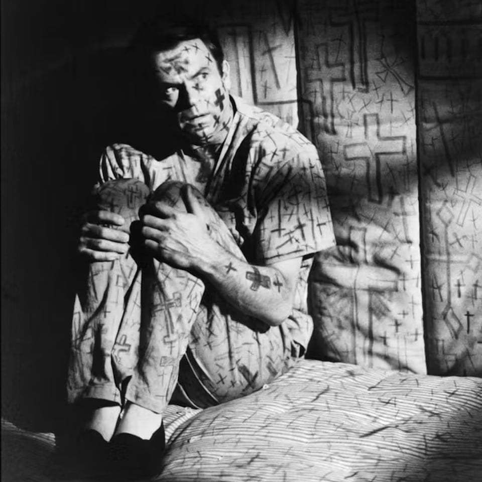 Sam Neill sits on the floor of his cell, looking fearful, clutching his knees. His face and clothes, and the walls and floor, are covered in hand-drawn crosses