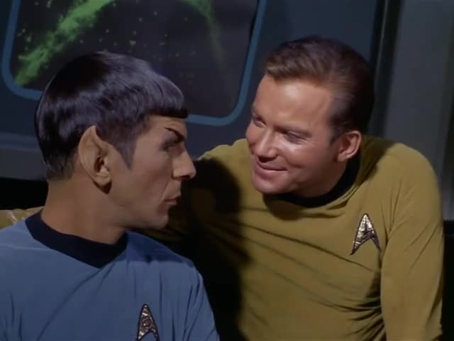 A still from Star Trek in which Captain Kirk is leaning over Spock with his arm around Spock, with a huge grin on Kirk's face