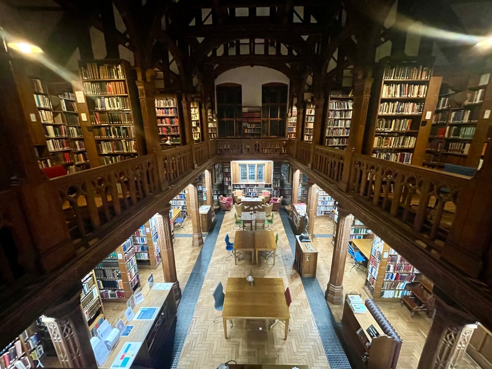 The photo is of Gladstone's Library, a two-floor library, taken from the top floor. The ground floor contains light-wood tables and chairs lined with bookcases. The top floor is mock-Tudor with dark-wood railings and rafters with bookcases along both sides.