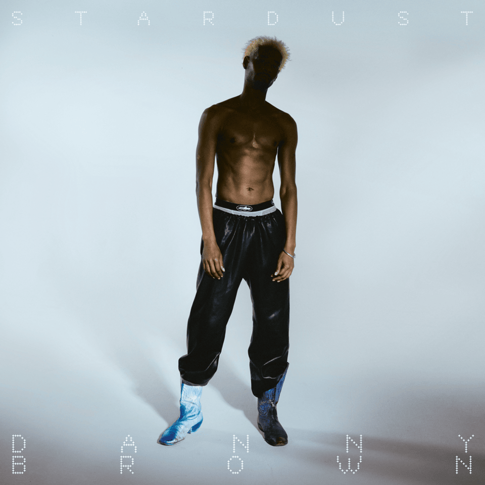 The cover of Danny Brown's album Stardust. The word 'stardust' is on top and the words 'danny brown' are on the bottom. He stands shirtless, in black reflective pants and galaxy-toned boots, with his face obscured by shadow.