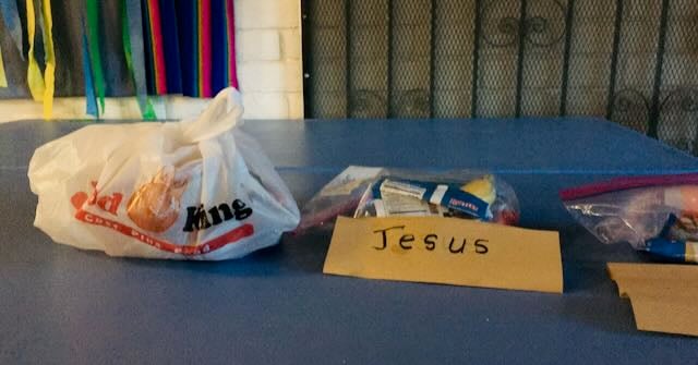 A table with a blue tablecloth and three bags of packaged snack food. The middle one has a paper label on it that says "Jesus."