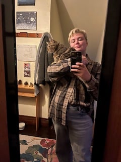 Nola in a mirror with a partially tucked in big button up and a cat on her shoulder