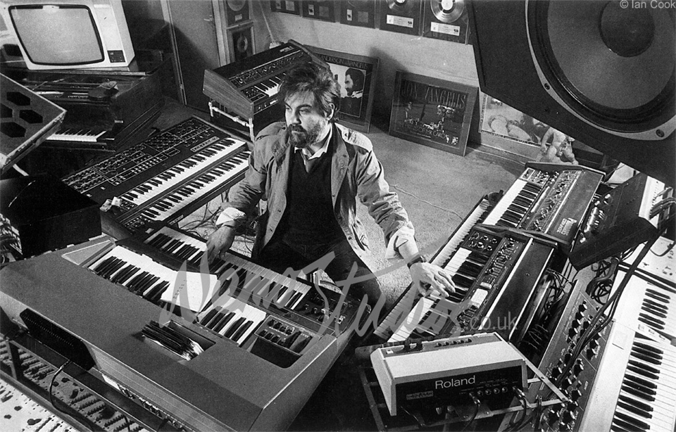 A black-and-white photo of the composer Vangelis, taken from above. He is sitting wearing a black sweater and a coat, and is sitting on a stool in a semicircle of large synthesizer and organs. He has his hand on two different organ keyboards