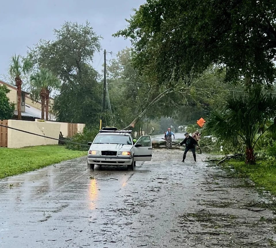 A residential street on a rainy day after a Hurricane.  A large tree fallen and blocks the entire street.  A power line is down on the side of the street.  Neighbors with chain saws are quickly cutting through the tree to clear it from the road.  A white Volvo station wagon is parked in front of the tree to pull away larger debris.  A neighbor dressed in black pants and a black jacket and hat is directing the car as it backs up to a large tree limb.