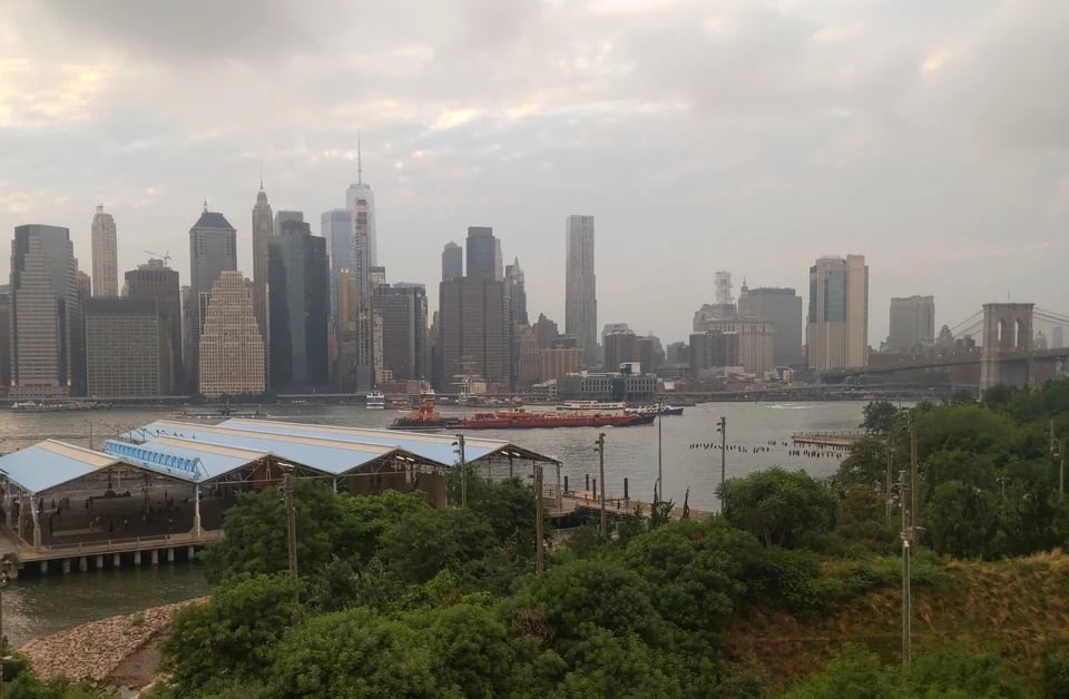 Photograph taken from the Brooklyn Heights Promenade on the evening of June 27, 2026. You can see a tug boat pushing a barge down the East River with the Manhattan Skyline in the background. It is a bit hazy.