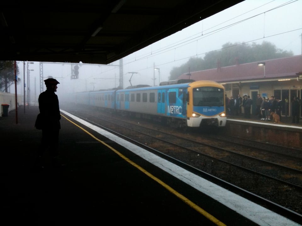 On the left hand side, a man, wearing what looks like a beret, is standing, waiting for a train at a train station. He looks to the right where a train is arriving at the station and the platform is full of people waiting to the get on the train. It's early morning and the whole scene is filled with fog, giving the whole a beautiful misty look.
