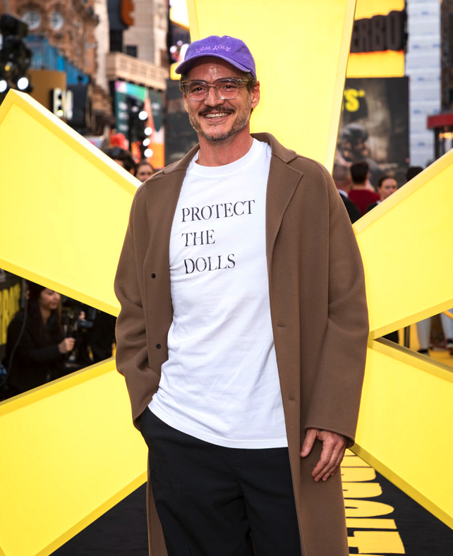 A scruffy Pedro Pascal in glasses, a long unbuttoned trench coat in dark camel, black slacks, and a white tee that says PROTECT THE DOLLS
