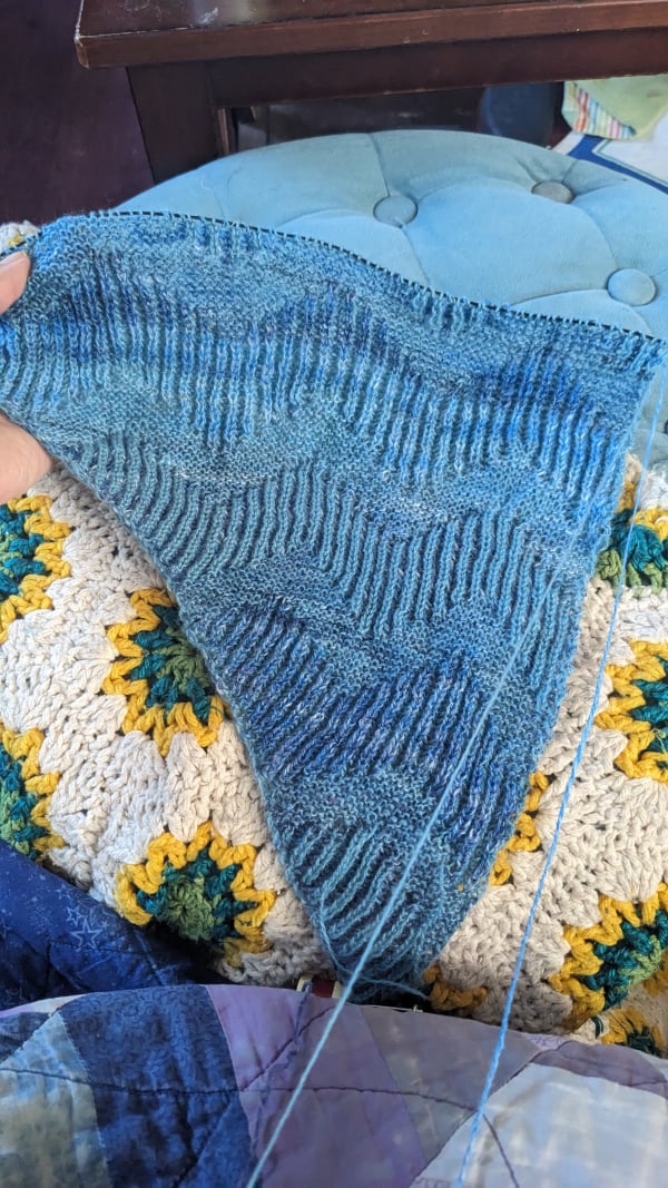 A triangular piece of knitting on the needles, in two shades of blue intertwined. Two different textures make stacks of alternating zigzag pattern.