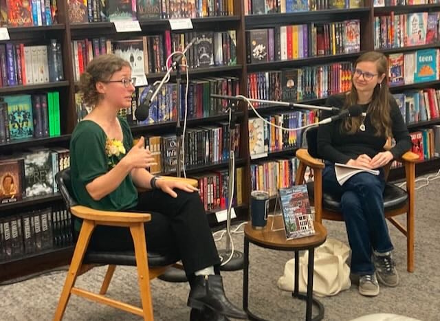 Two women are seated in a bookstore, engaged in conversation during an event, with bookshelves filled with colorful books in the background.
