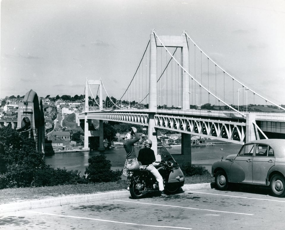 Two bridges crossing a river, with a town on the other side. To the left is a narrow railway bridge, low dark and heavy. To the right is a new road suspension bridge, looking light and modern. In the foreground, a man is parked on a scooter, with a woman sitting pillion behind him.