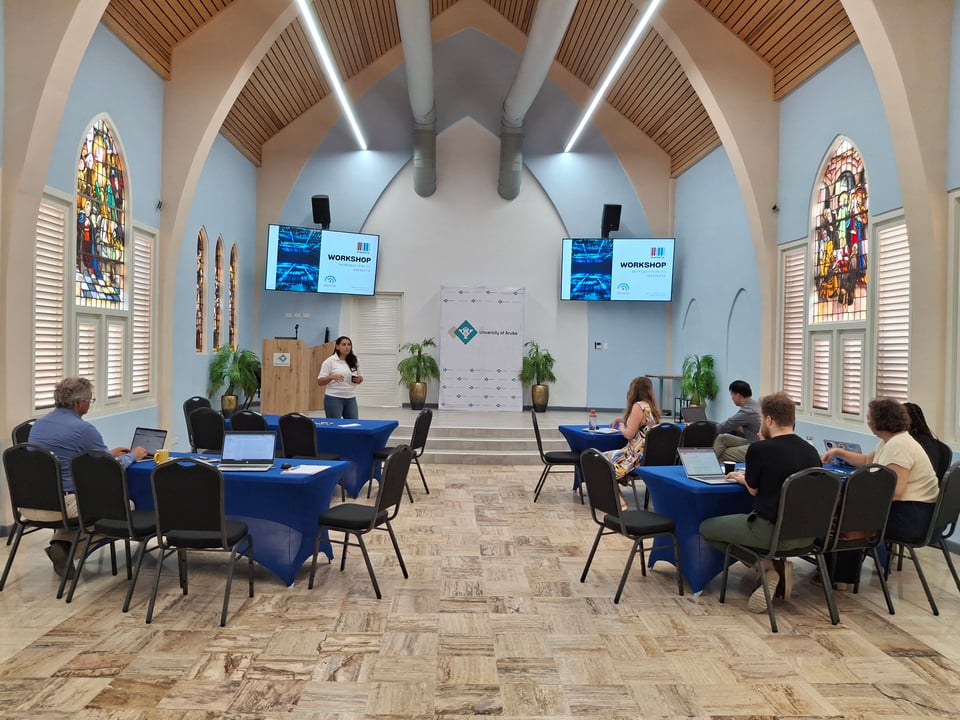 A picture of the wikidata workshop in progress, with Ginelly in the front of the MC Chapel at the university of Aruba, presenting on Wikidata. The workshop participants are seated around tables and have their laptop open, waiting for the practical part of the workshop where they will add information to wikidata.