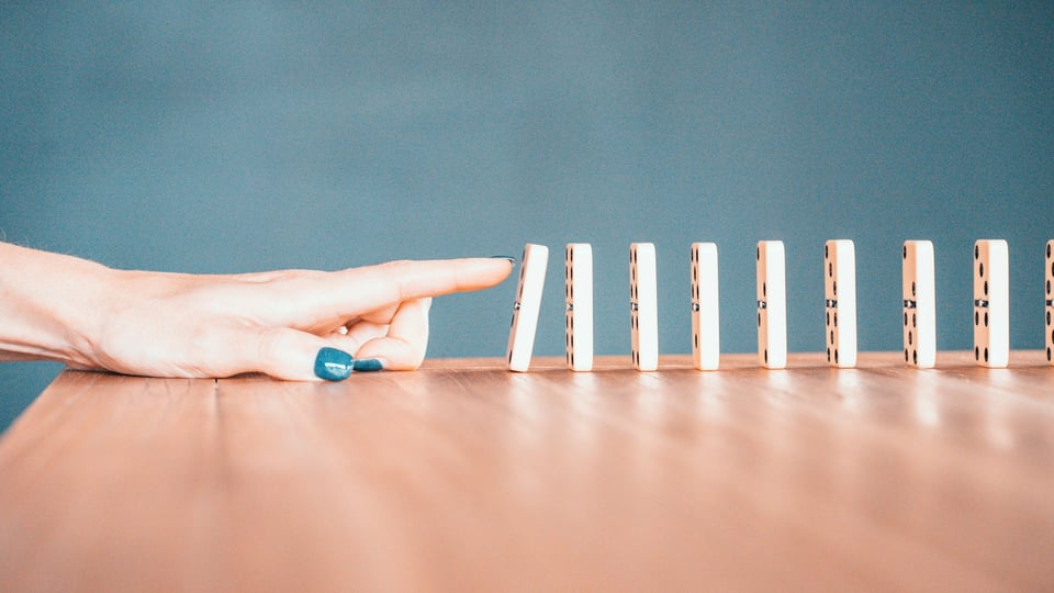 A finger tipping over the first in a row of dominoes. Photo by Bradyn Trollip on Unsplash.