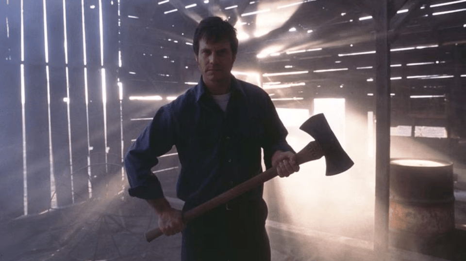 A still from the movie Frailty, featuring Bill Paxton wielding an axe in a rickety barn backlit by ominous white light.