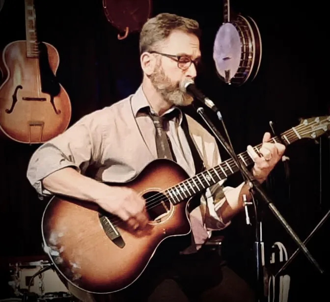 A man with a beard and glasses wearing a button-down shirt with a loosened necktie plays an acoustic guitar and sings into a microphone.