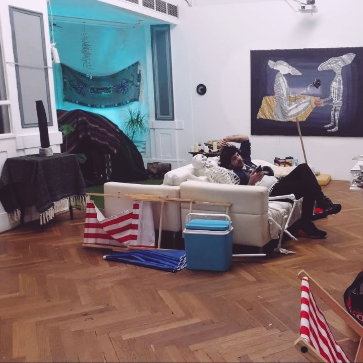 A photo of the artist sitting on a sofa next to a skeleton, surrounded by artworks on the walls and a tent behind him.