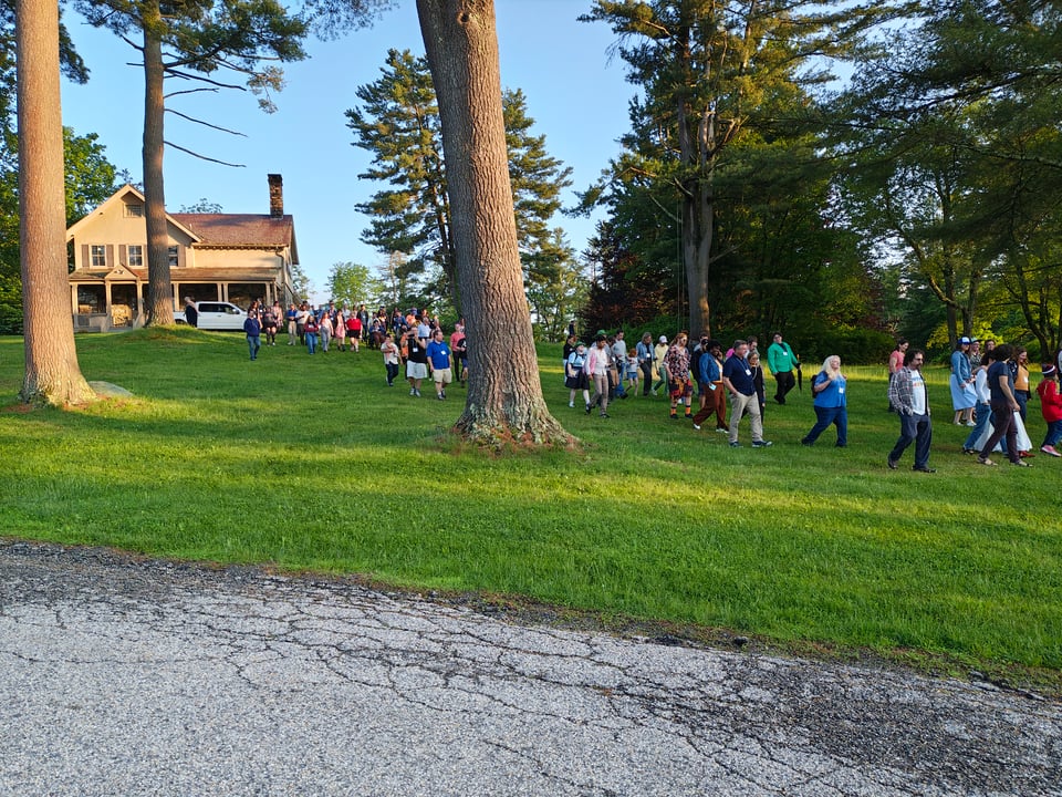Several dozen people in casual clothes make their way down a gentle hill from an old stone house across a vibrant green lawn and past stately oak and pine trees toward a roughly paved road in the foreground