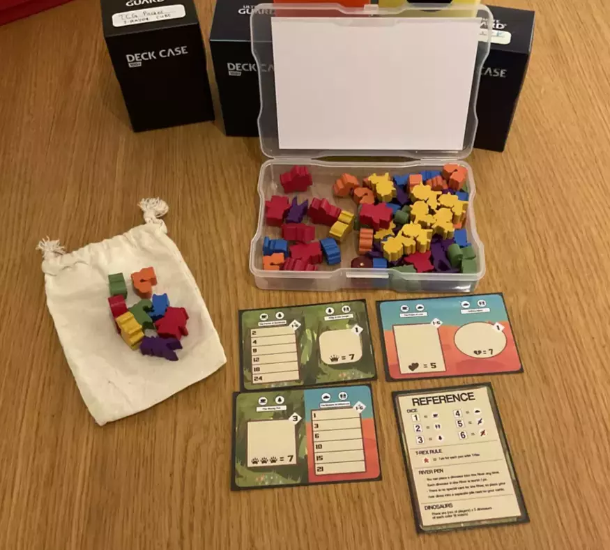 A clear plastic case full of colorful wooden meeples. In front of the box, a small cloth pouch with more meeples on top and four cards depicting different dinosaur pens and a reference card.
