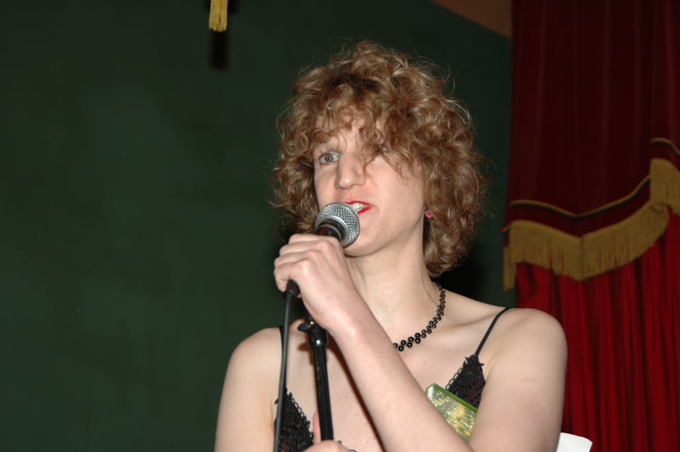A white trans woman with curly brown hair wearing a strappy black dress and black necklace, holding a microphone with a notebook under one arm. Behind her is a green wall and a red curtain.